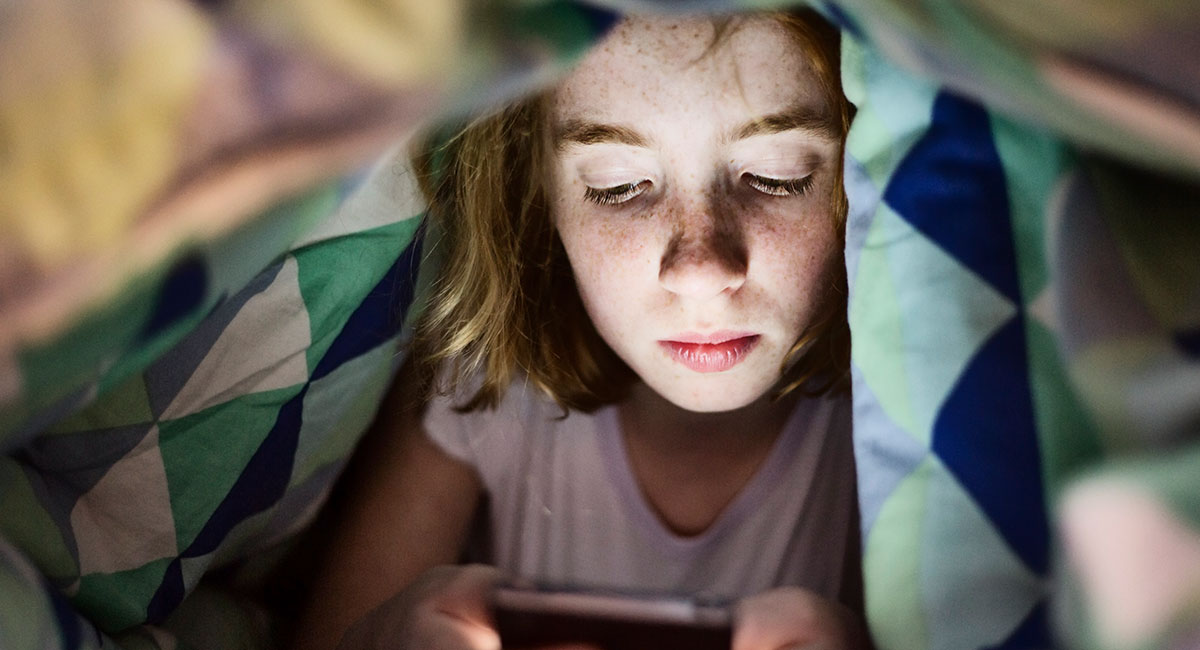 Flicka i yngre tonåren tittar på sin smartphoneskärm under ett täcke.