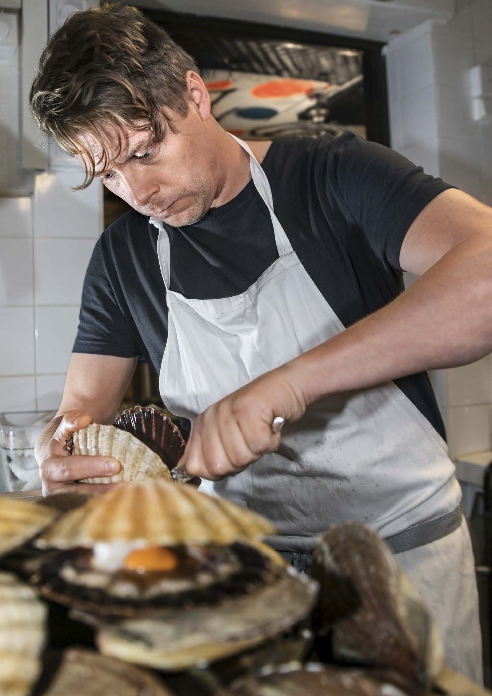 Joakim har på sig förkläde och öppnar kammusslor i restaurangmiljö