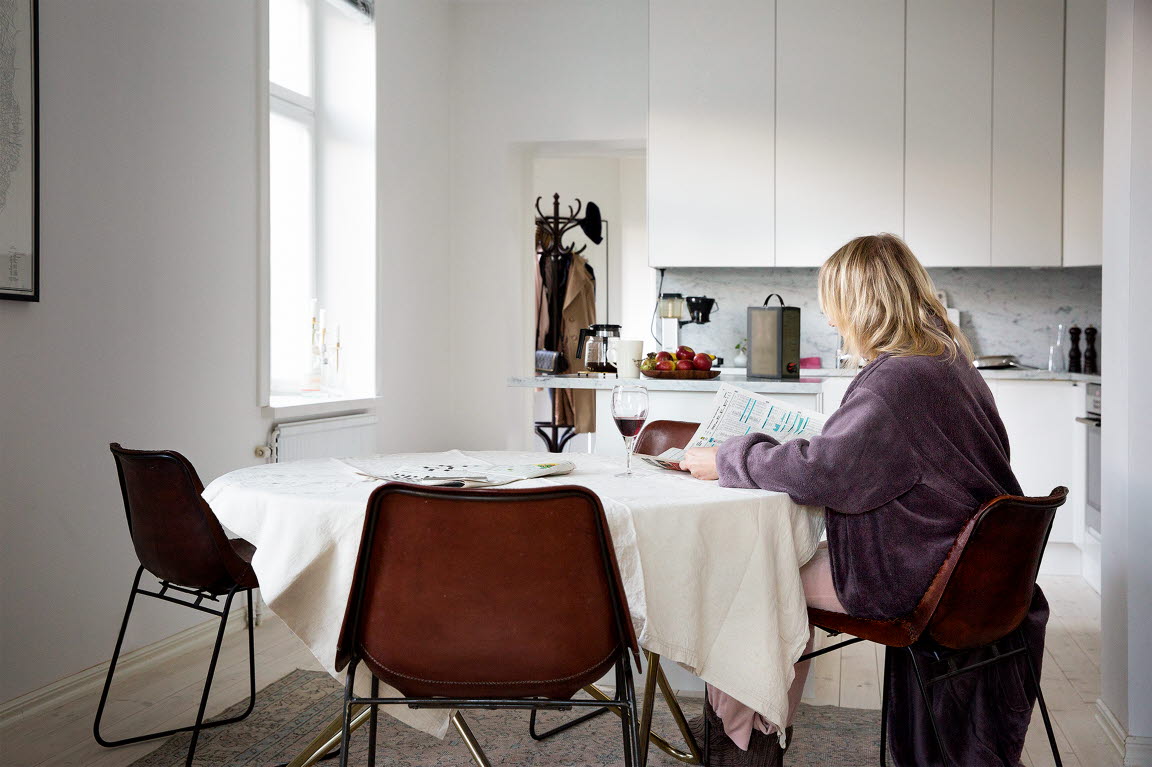 En kvinna i morgonrock sitter vid köksbordet. Hon läser tidningen och på bordet står ett glad vin.