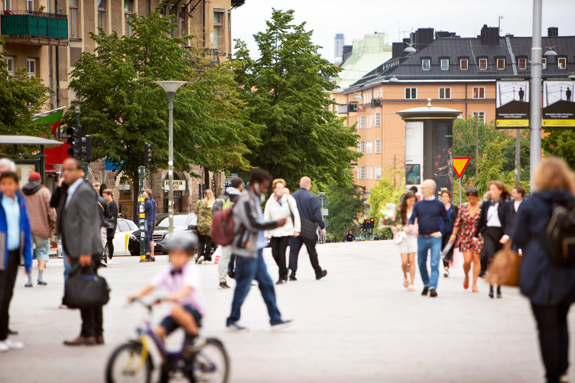 Bild tagen på vimlet av människor på en gågata i Stockholm. Vi ser bland annat en liten pojke på en cykel, en man som håller i en portfölj och en kvinna i blommig klänning. Det är sommar och fint väder.