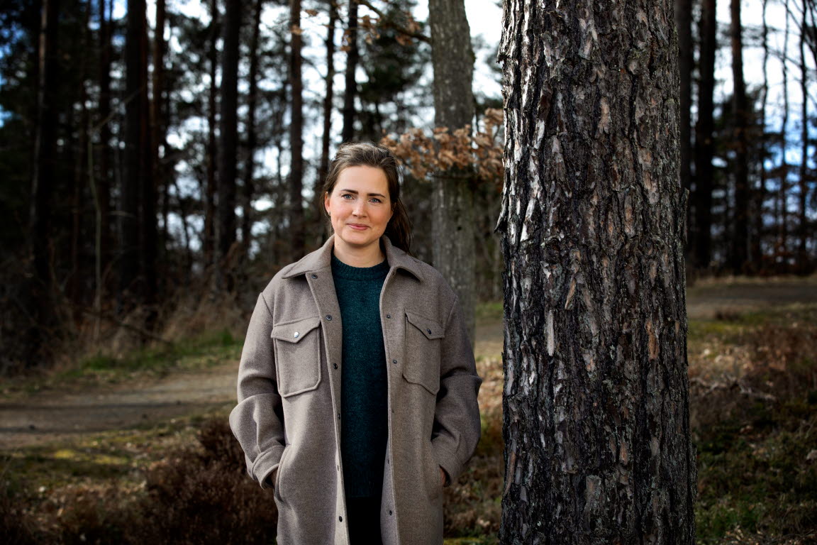  	Felicia Ahlner står i en skog bredvid ett träd. Hon ler in i kameran.