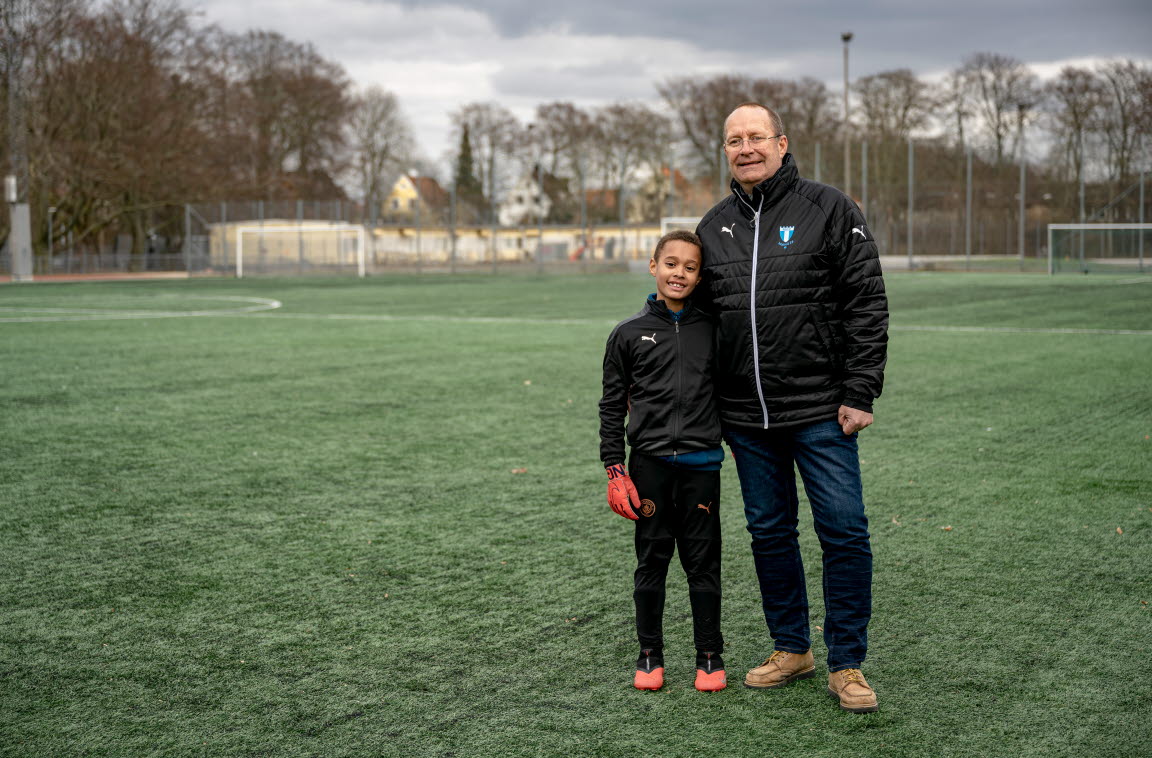 Mats Magnusson står på en fotbollsplan med armen om hans son. Sonen har på sig träningskläder och Mats har på sig en Malmö FF-jacka