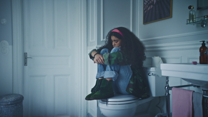 Ung tjej sitter med bena uppdragna mot sig på en toalett. Hon ser nedstämd ut.