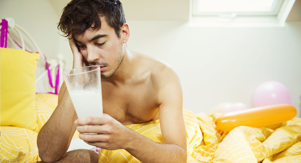 Bakfull man sitter i sängen och mår dåligt. Han håller i ett stort glas innehållandes huvudvärksmedicin upplöst i vatten.