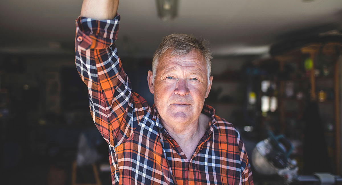 Äldre man i rutig skjorta befinner sig i ett garage. Han tittar in i kameran och ser neutralt in i kameran