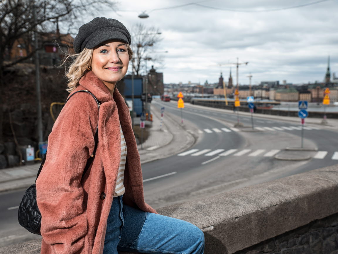 Vanja Wikström iklädd en mörkrosa kappa sitter på en mur bredvid en väg. Hon tittar leende in i kameran