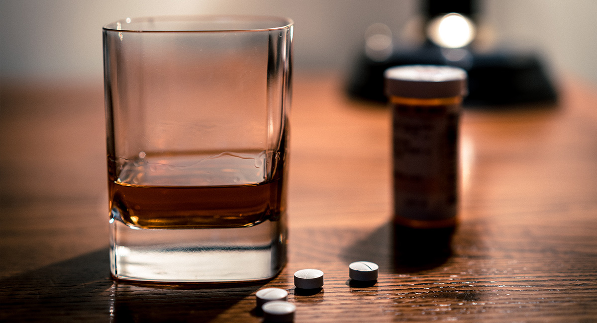 Närbild på ett glas med whiskey som står på ett bord, bredvid ligger några piller och en pillerburk