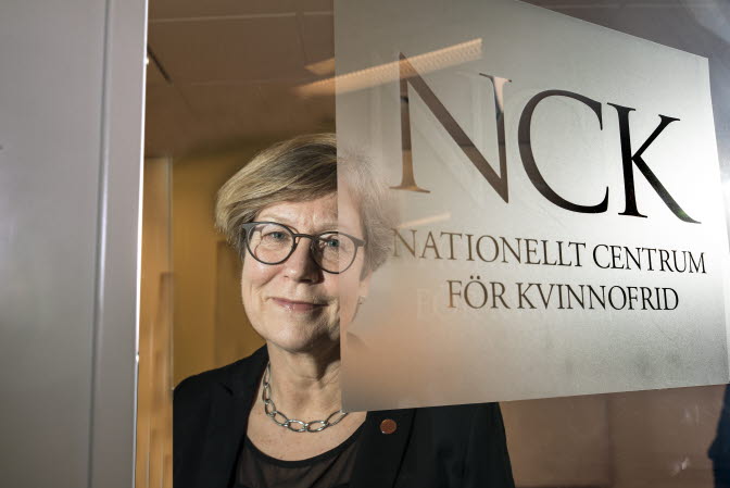Åsa Witkowski står bakom en glasdörr med NCK:s logga. Hon tittar leende in i kameran och har på sig glasögon.